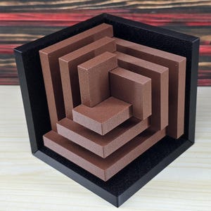 May include: A three-dimensional geometric sculpture featuring a series of nested, brown rectangular bars arranged in a spiral pattern. The structure is encased within a black, open-faced cube frame, creating a layered, architectural appearance.