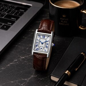 May include: A rectangular wristwatch with a silver-tone case, a white dial featuring blue Roman numeral hour markers, and a brown crocodile-embossed leather strap. The watch has blue hands and a small sub-dial at the bottom of the face.