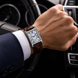 May include: A rectangular wristwatch with a silver-tone case, a white dial featuring black Roman numerals, and blue hands. The watch is secured with a dark brown textured leather strap with a visible grain pattern and a polished metal buckle.