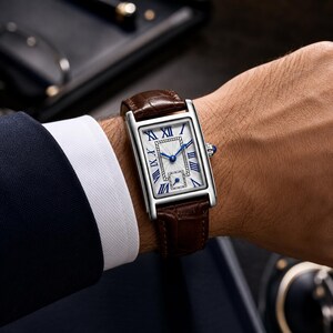 May include: A rectangular wristwatch with a silver-toned case and a brown alligator-textured leather strap. The white dial features blue Roman numeral hour markers, blue hands, a central textured rectangular pattern, and a small sub-dial at the bottom.
