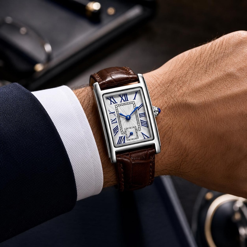 May include: A rectangular wristwatch with a silver-toned case and a brown alligator-textured leather strap. The white dial features blue Roman numeral hour markers, blue hands, a central textured rectangular pattern, and a small sub-dial at the bottom.