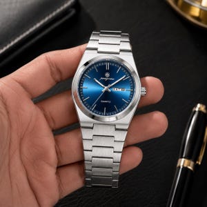 May include: A silver-toned stainless steel wristwatch featuring a circular deep blue sunray dial with silver hour markers and hands. The watch face displays the text "paagomu" and "QUARTZ" with a day-date window at the three o'clock position.
