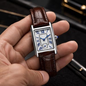 May include: A rectangular wristwatch with a silver-tone case and a dark brown embossed leather strap. The white dial features blue Roman numeral hour markers, blue hands, a central textured rectangular pattern, and a small sub-dial at the bottom.