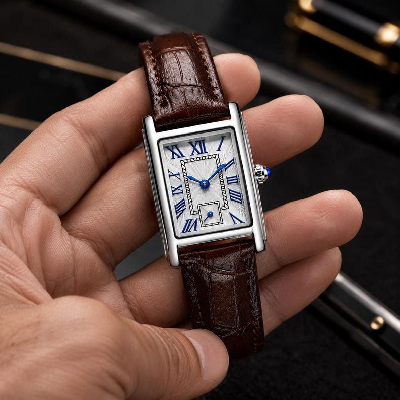 May include: A rectangular wristwatch with a silver-tone case and a dark brown embossed leather strap. The white dial features blue Roman numeral hour markers, blue hands, a central textured rectangular pattern, and a small sub-dial at the bottom.