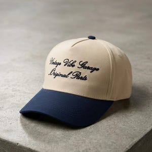 Pode incluir: Boné de beisebol bege com aba e botão azul marinho. O boné apresenta o texto bordado "Vintage Vibe Garage Original Parts" em azul marinho. Feito de tecido durável, é adequado para uso casual.
