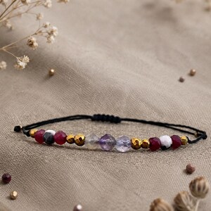 Può includere: Bracciale fatto a mano con cordino nero e chiusura a nodo scorrevole. È adornato con perline dorate, pietre preziose rosse, bianche e viola. Il bracciale è presentato su una superficie neutra. Lunghezza regolabile.