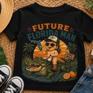 May include: Black t-shirt with a graphic of a baby riding an alligator, sunglasses, and the text "FUTURE FLORIDA MAN." The design includes palm trees, an orange sunset, and the words "STAY WILD."