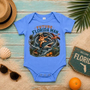 May include: A light blue baby bodysuit featuring a graphic print of a baby wearing sunglasses and a cap, riding a shark in a swirling ocean wave. The text reads FUTURE FLORIDA MAN. The design includes a flamingo, an alligator, and oranges.