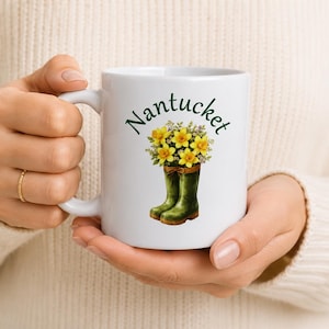 Pode incluir: Caneca de cerâmica branca com a palavra "Nantucket" em escrita verde. A caneca apresenta uma ilustração em aquarela de flores amarelas em galochas verdes. A caneca é segurada por uma pessoa que usa um suéter creme.