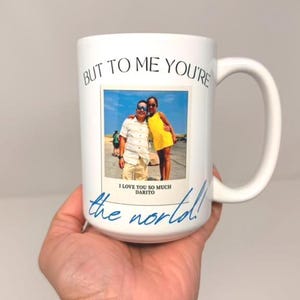 Pode incluir: Uma caneca de cerâmica branca com uma fotografia impressa de duas pessoas em pé ao ar livre. O texto "BUT TO ME YOU'RE" está impresso a preto acima da foto, e "the world!" está escrito em azul abaixo da foto com "I LOVE YOU SO MUCH DARITO".