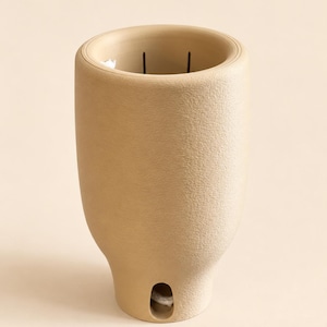 May include: A beige, textured, cylindrical vessel with a rounded top rim and a small, oval-shaped opening near the base. The interior of the vessel is hollow, featuring a visible wick, suggesting it is a candle container or a planter.
