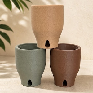 Può includere: Tre vasi cilindrici con finitura opaca testurizzata, impilati a piramide. Il vaso superiore è beige, quello in basso a sinistra è verde salvia e quello in basso a destra è marrone. Ogni vaso presenta un foro di drenaggio a forma di goccia.