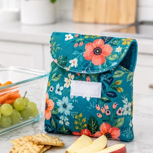 Puede incluir: Una bolsa para snacks de tela color verde azulado con un estampado floral vibrante de flores color coral, amarillas y blancas con hojas verdes. La bolsa tiene una solapa plegable asegurada con un cierre de velcro blanco rectangular al frente.