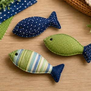 Può includere: Tre giocattoli a forma di pesce in tessuto imbottito. Uno è blu scuro con pois bianchi. Un altro è verde tinta unita con una finitura testurizzata. Il terzo presenta strisce verticali verdi, blu e bianche con una coda blu scuro. Ogni pesce ha un piccolo occhio nero.