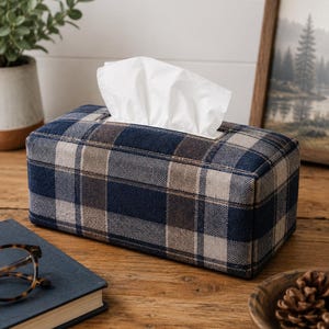 May include: A rectangular tissue box cover featuring a woven fabric pattern in navy blue, tan, and dark brown plaid. The cover is designed to fit over a standard rectangular tissue box, with a white tissue protruding from the top opening.