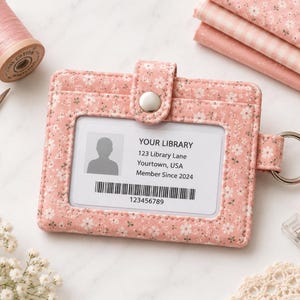 May include: A pink fabric ID card holder with a small white floral pattern, featuring a snap button closure and a metal ring. The holder contains a library card with the text YOUR LIBRARY, 123 Library Lane, Yourtown, USA, Member Since 2024.