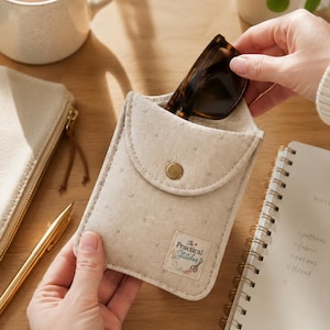 May include: A beige felt glasses case with a snap closure, holding a pair of tortoiseshell sunglasses. The case has a small label that reads "The Practical Stitches". A gold pen and a notebook are also visible.