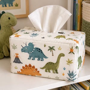 May include: A rectangular fabric tissue box cover featuring a repeating pattern of various dinosaurs, palm trees, and volcanoes in shades of blue, green, and orange on an off-white background, with a single white tissue pulled through the top slot.