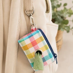 Puede incluir: Un dispensador de bolsas para excrementos de perro a cuadros multicolores con cremallera azul y clip plateado. El dispensador está sujeto a una correa de bolso beige y se ve una bolsa de residuos verde.