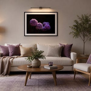 Peut inclure: Une impression encadrée d'art floral représentant des fleurs d'ail pourpre sur fond sombre. Le tableau est au-dessus d'un canapé blanc avec des coussins et une couverture. Une table basse ronde en bois est devant le canapé.