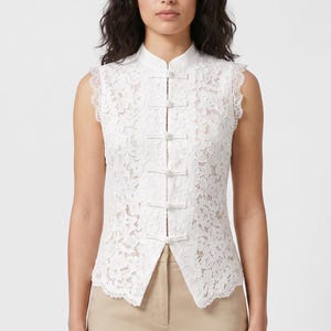 May include: A sleeveless white lace top featuring a mandarin collar and a front closure with five fabric-covered toggle buttons. The garment is constructed from a floral lace pattern with a scalloped hemline and is paired with tan trousers.