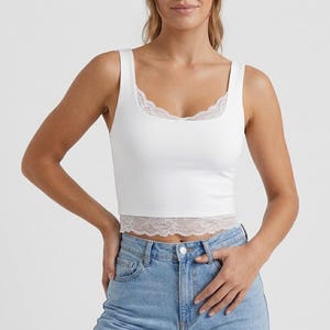 May include: White tank top with a square neckline and lace trim along the neckline and hem. The top is paired with light blue denim jeans. The overall style is casual and modern.