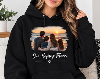 May include: A black hooded sweatshirt featuring a rectangular graphic print of a family of four sitting on a beach at sunset. Below the image, the text reads Our Happy Place in white script, accompanied by a small white heart and horizontal lines.