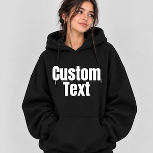May include: A black hooded sweatshirt featuring the words Custom Text printed in large white capital letters across the chest. The garment includes a front kangaroo pocket, long sleeves with ribbed cuffs, and a drawstring hood with matching black cords.