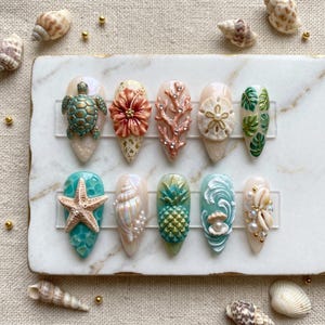 May include: A set of ten artificial nails with a beach theme. Each nail features a different design, including a sea turtle, coral, starfish, seashell, pineapple, and tropical leaves. The nails are painted in various colors, including teal, gold, and white.