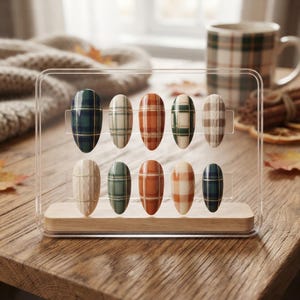 May include: A set of ten almond-shaped press-on nails displayed on a clear acrylic stand with a wooden base. The nails feature various plaid and cable knit patterns in shades of forest green, burnt orange, cream, and brown with gold line accents.