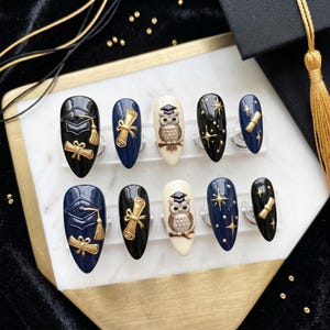 May include: A set of ten graduation-themed false nails. Designs include a mortarboard, diploma, owl, and stars in navy, black, and cream colours. Gold accents are used throughout the nail art.