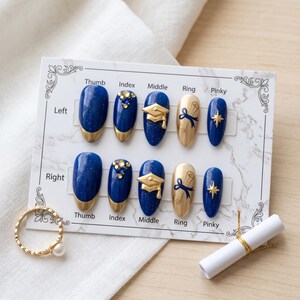 May include: A set of press-on nails with a graduation theme. The nails feature a navy blue base with gold accents, including a gold French tip, graduation caps, and scroll designs. A gold ring with a pearl and a rolled diploma are also in the image.