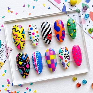 Puede incluir: Un juego de diez uñas postizas en forma de almendra sobre un soporte de acrílico transparente. Cada uña presenta un patrón distinto inspirado en los años 90, incluyendo garabatos amarillo neón, puntos coloridos, rayas blancas y negras, cuadros y formas geométricas en tonos vibrantes de rosa, azul, naranja, verde y negro.