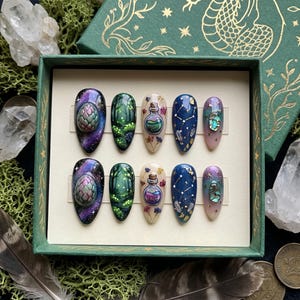 Può includere: Un set di dieci unghie finte con vari disegni. I disegni includono una galassia, una scena di foresta, una bottiglia di pozione, una notte stellata e una conchiglia di abalone. Le unghie sono presentate in una scatola verde con dettagli dorati.