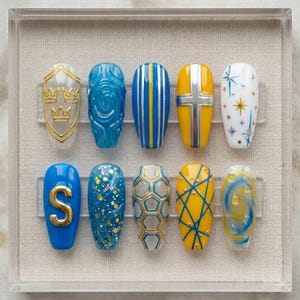 Può includere: Un set di dieci unghie finte con vari disegni. Le unghie presentano una gamma di colori tra cui blu, giallo e bianco, con accenti dorati. I disegni includono corone, vortici, strisce, croci, stelle, la lettera "S", glitter e motivi geometrici.