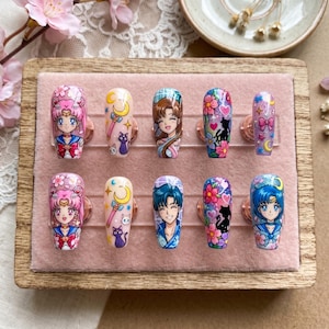 May include: A set of ten press-on nails with anime-style character designs. Each nail features a different design, including characters, moons, stars, and floral patterns. The nails are displayed on a pink velvet surface.