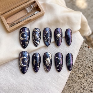 Puede incluir: Un juego de diez uñas postizas en forma de almendra con un acabado de galaxia en azul y morado profundo, con detalles celestiales metálicos en plata que incluyen lunas, estrellas, constelaciones y pequeños diamantes de imitación, con un kit de madera.