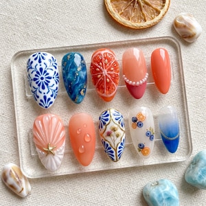 Pode incluir: Um conjunto de dez unhas artificiais com vários designs. Os designs incluem padrões florais, efeitos de mármore, fatias de frutas e gotas de água. As cores incluem azul, laranja, branco e rosa. Algumas unhas apresentam pérolas e detalhes dourados.