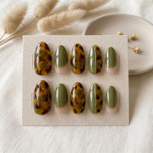 May include: A set of ten press-on nails arranged on a rectangular card. The nails feature a mix of glossy tortoise shell patterns in brown and green, solid olive green, and olive green with a vertical gold line accent, all with a rounded shape.