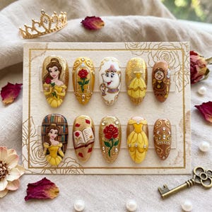 Könnte beinhalten: Ein Set aus zehn künstlichen Nägeln mit Designs zum Thema Die Schöne und das Biest. Die Nägel zeigen Charaktere wie Belle, Madame Pottine und Lumière, zusammen mit Rosen und Kleidern. Die Farbpalette umfasst Gold, Gelb und Braun.