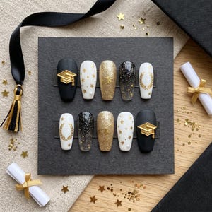 May include: A set of ten press-on nails in various designs. The nails feature black, white, and gold colours with graduation cap, star, and laurel leaf accents. Some nails have glitter effects, and the set is arranged on a black card.