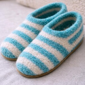 May include: A pair of slip-on slippers featuring a fuzzy, textured surface with horizontal stripes in alternating shades of bright turquoise blue and off-white. The slippers have a rounded toe design and a solid tan-colored flat sole base.