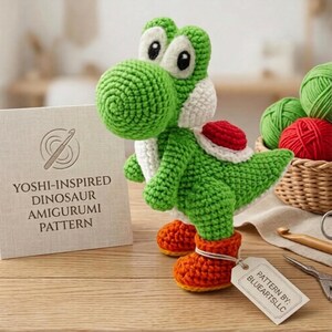 Könnte beinhalten: Ein grünes, weißes und rotes gehäkeltes Yoshi-Dinosaurier-Amigurumi steht auf einer Holzoberfläche. Die handgefertigte Figur hat orangefarbene Stiefel und große, weiß umrandete Augen. Ein Etikett lautet "Pattern by: BLUEARTSLLC". Ein Schild lautet "Yoshi-Inspired Dinosaur Amigurumi Pattern".