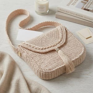 Puede incluir: Bolso bandolera de crochet beige con cierre de solapa y correa larga. El bolso presenta una borla decorativa y un pequeño detalle metálico. Hecho de hilo texturizado, está sobre una superficie blanca.