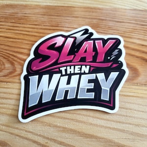 Może przedstawiać: Naklejka z napisem "SLAY THEN WHEY". Słowo "SLAY" jest w kolorze różowym z czarnym obrysem, a "WHEY" jest białe z szarym i czarnym obrysem. Naklejka znajduje się na drewnianej powierzchni.