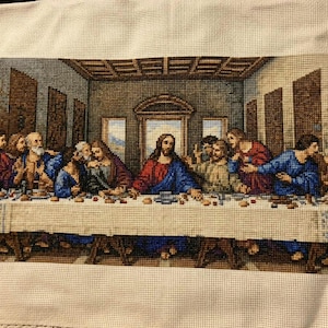 Può includere: Un ricamo a punto croce che raffigura L'Ultima Cena, con una figura centrale in tunica rossa e blu seduta a un lungo tavolo con dodici figure. La scena è ambientata in una stanza con dettagli architettonici, realizzata con fili di vari colori su un tessuto di colore chiaro.