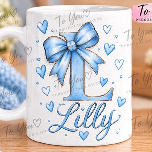 May include: A white ceramic mug featuring a large blue letter L adorned with a blue ribbon bow at the top. The design includes scattered blue hearts and the name Lilly written in blue cursive script below the letter. The mug has a handle on the left.