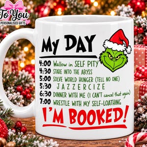 May include: A white ceramic mug featuring a green cartoon character wearing a Santa hat and a schedule titled MY DAY. The text lists activities including wallow in self pity, stare into the abyss, solve world hunger, jazzercize, and I'M BOOKED!