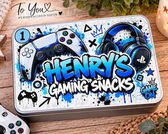 May include: A rectangular metal tin featuring a white background with blue paint splatter graphics. The lid displays a white and black game controller, blue headphones, and the text Henry's Gaming Snacks in bold blue and white lettering.