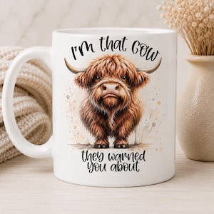 May include: A white ceramic mug featuring a brown Highland cow illustration centred on the front. Above the cow, the text reads I'm that cow, and below the cow, the text reads they warned you about. The mug has a curved handle on the left side.